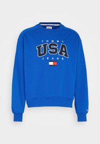 Felpa blu con scollo rotondo, che presenta "TOMMY USA JEANS" in grassetto bianco e nero, e un piccolo logo di bandiera come accento.