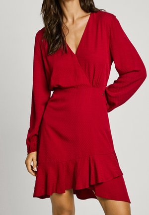 Kobieta ubrana w czerwoną sukienkę kopertową z długim rękawem, z falbaniastym asymetrycznym dołem, teksturowaną tkaniną i dekoltem w kształcie litery V.