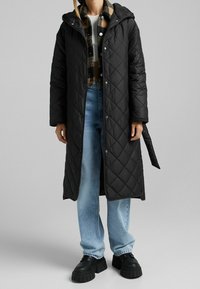 Długi czarny pikowany płaszcz z kapturem, z zatrzaskami, noszony na koszuli w kratę oraz w połączeniu z jasnoniebieskimi dżinsami i masywnymi czarnymi butami.