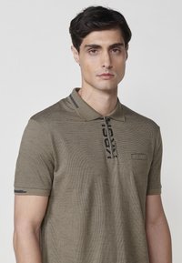 Camisa polo caqui de manga corta, fabricada en un tejido texturizado, con un cuello con cremallera y un detalle de logo negro que recorre verticalmente la parte delantera.