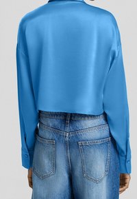 Persona che indossa una camicia a maniche lunghe di un brillante blu, accorciata e abbinata a jeans a vita alta di un blu sbiadito, vista da dietro.