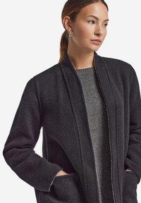 Svart ullblandad kappa med öppet framdesign, sidofickor och en texturerad yta, lagd över en grå stickad tröja.