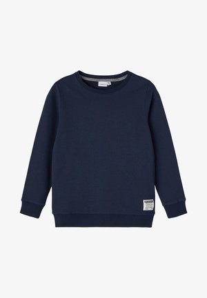 Granatowa bluza z długim rękawem i okrągłym dekoltem, z prążkowanymi mankietami i ściągaczem na dole, z małą białą naszywką z tekstem blisko dolnego brzegu.