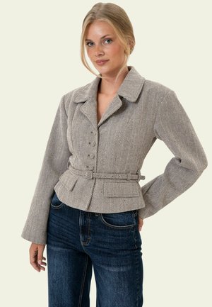 Femme blonde portant un blazer beige à motif chevrons avec une ceinture à la taille et un jean bleu foncé, debout avec une main sur la hanche.