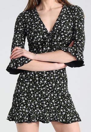Woman wearing a black dress with white floral print, V-neck, three-quarter ruffled sleeves, and a ruffled hem, arms crossed at waist.