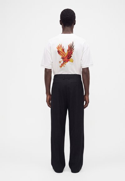 Ein Mann steht mit dem Rücken zur Kamera, trägt ein weißes T-Shirt mit einer rot-gelben Adlergrafik auf dem Rücken und schwarze, weit geschnittene Hosen.