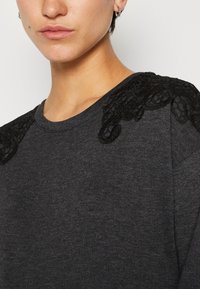 Sweatshirt gris foncé avec des détails en dentelle noire texturée sur les épaules et un col rond. Tissu doux avec une coupe décontractée.