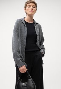 Camicia a righe bianca e nera con bottoni, abbinata a un top nero aderente e pantaloni neri a vita alta; tiene in mano una borsa nera strutturata.