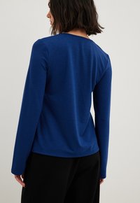 Frau von hinten gesehen, die ein langärmliges blaues Oberteil und schwarze Hosen trägt, vor einem schlichten hellen Hintergrund.