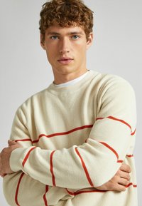 Kreminės spalvos megztinis su raudonomis horizontaliomis juostelėmis, gofruota tekstūra, apvalus iškirpimas ir ilgomis rankovėmis. Audinys atrodo minkštas ir pralaidus orui.