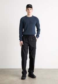 Giovane uomo in piedi, vestito con un maglione di lana blu navy, pantaloni cargo neri, sneakers nere e un cappellino nero con un piccolo logo.
