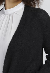 Mustavilla neulecardigan, joka on pehmeä kosketukseltaan, avoin edestä ja leveällä kauluksella, yhdistetty valkoisen pliseeratun paidan kanssa.