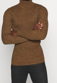 Personne portant un pull à col roulé marron tacheté, les bras croisés, associée à un pantalon noir, sur un fond clair uni.