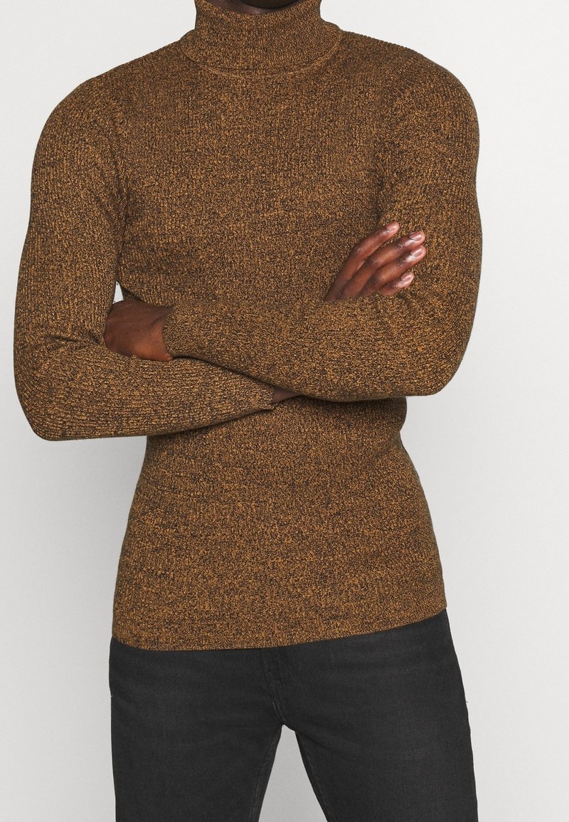 Personne portant un pull à col roulé marron tacheté, les bras croisés, associée à un pantalon noir, sur un fond clair uni.