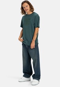 Grünes T-Shirt mit kurzen Ärmeln und dunklem Logo, kombiniert mit lässigen blauen Jeans und weißen Sneakern auf einem einfarbigen Hintergrund.