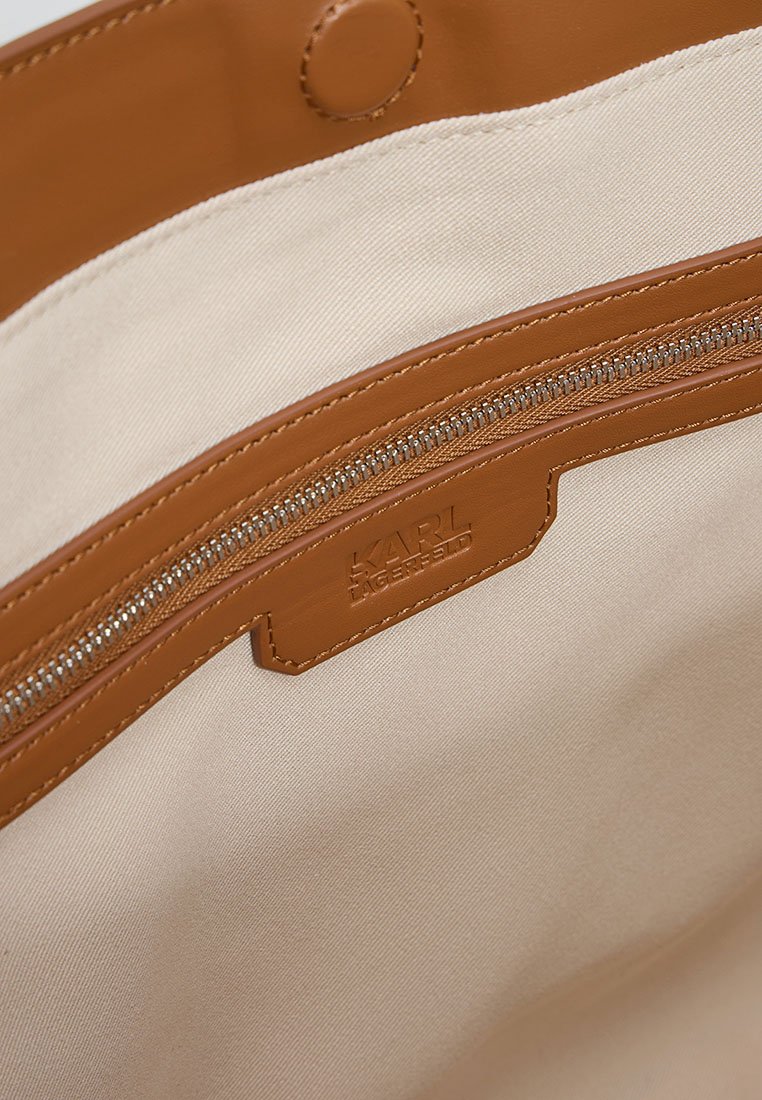 Braune Lederhandtasche mit cremefarbener Stoffauskleidung. Verfügt über ein Reißverschlussfach und einen geprägten Logo-Bereich. Glatte Textur und sauberes Design.