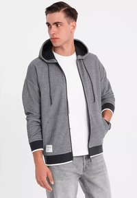 Sudadera gris con cremallera y acentos negros, cordones y bolsillos laterales. Lleva puesta sobre una camiseta blanca, combinada con jeans gris claro.