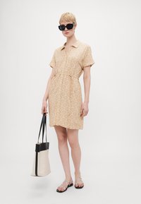 Light beige dress with shell pattern; short sleeves; collar; drawstring waist. Model holds a black and beige tote bag, wearing black sunglasses.