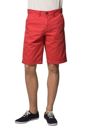 Homme portant un short rouge mi-genoux, une chemise blanche rentrée et des baskets noires à lacets avec des semelles blanches.