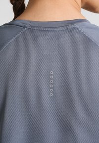 Camiseta deportiva gris con tela de malla texturizada. Presenta un cuello redondo y una fila vertical de pequeños botones en la parte trasera.