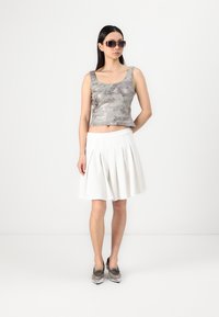 Ezüst fémes crop top, fehér pliszírozott szoknya és ezüst hegyes cipők. A modellt nagy napszemüvegben láthatjuk, egy semleges háttér előtt.