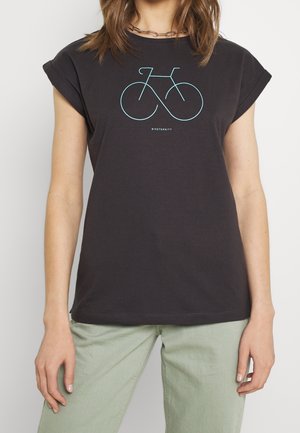 Frau trägt ein schwarzes T-Shirt mit türkisfarbener Linienzeichnung eines Fahrrads und dem Text "BIKETERNTIY", kombiniert mit hellgrünen Hosen vor einfarbigem Hintergrund.