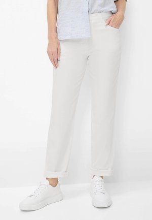 Person trägt hellgraue, hochgekrempelte Hose, ein hellblau gemustertes Hemd, weiße Turnschuhe, mit einer Hand in der Tasche und der anderen am Bein.