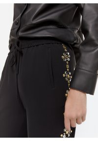 Close-up van een hand naast zwarte broek met gouden en zilveren edelsteenversieringen, gecombineerd met de mouw van een zwarte leren jas.