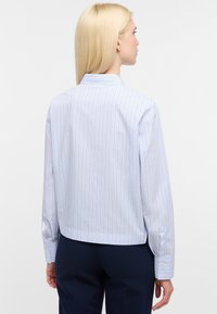 Donna con capelli biondi lisci, indossa una camicia a righe blu chiaro con maniche lunghe e pantaloni scuri, vista di spalle su uno sfondo neutro.