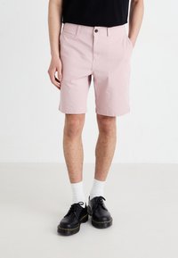 Shorts rose clair en coton, avec une fermeture à bouton, des poches latérales et des ourlets retroussés. Portés avec des chaussures noires et des chaussettes blanches.