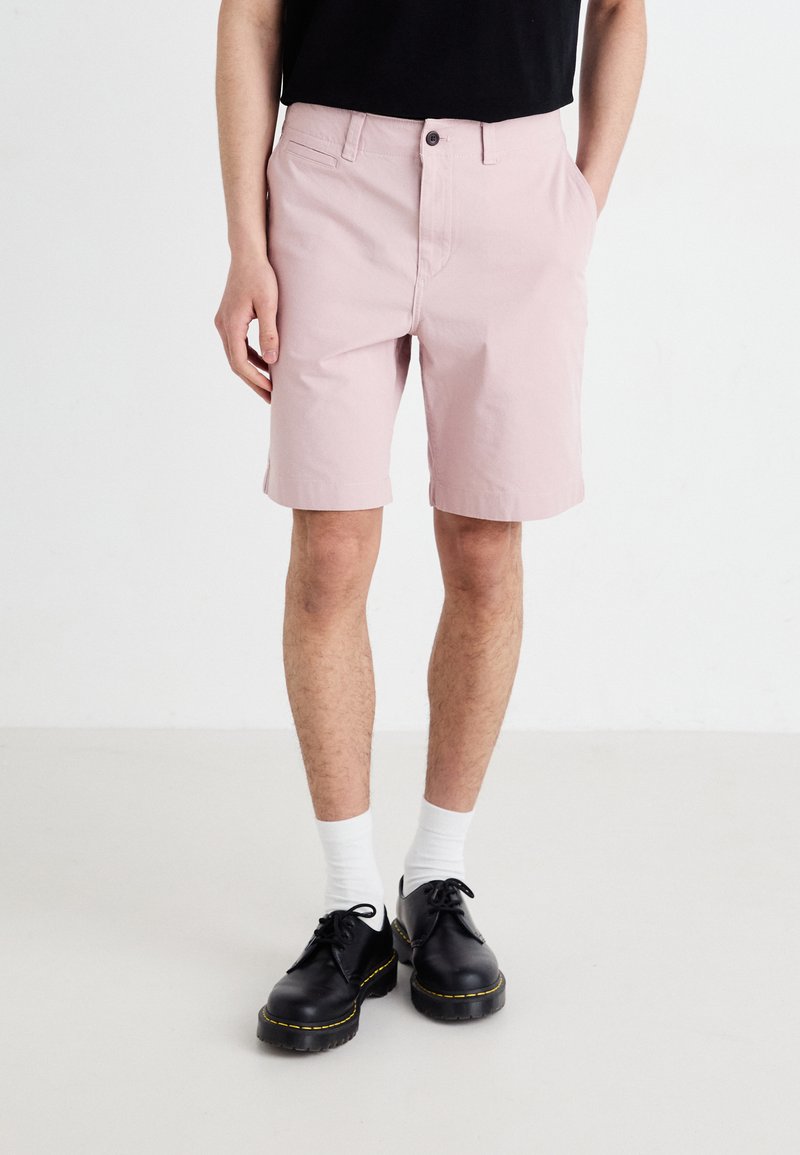 Shorts rose clair en coton, avec une fermeture à bouton, des poches latérales et des ourlets retroussés. Portés avec des chaussures noires et des chaussettes blanches.