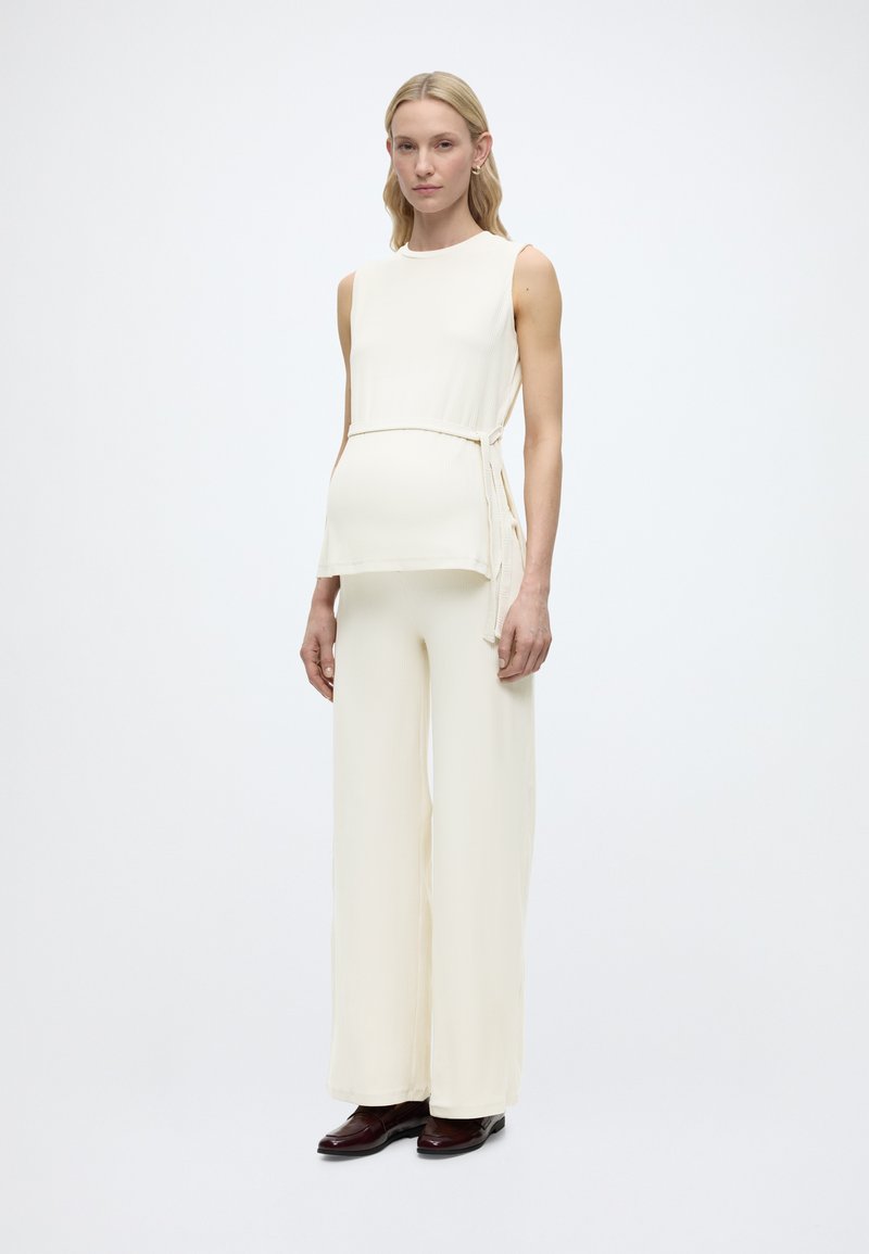 Γυναίκα στέκεται φορώντας αμάνικη κρεμ τοπ με δεσίματα στο πλάι και αντίστοιχο φαρδύ παντελόνι, σε συνδυασμό με σκούρα καφέ παπούτσια, σε λευκό φόντο.