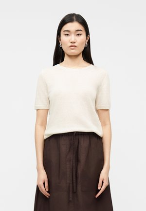 Žena s dlhými tmavými vlasmi, ktorá má na sebe strieborné kruhové náušnice, krémový krátky sveter s rukávmi a tmavohnedú sukňu s vysokým pásom a šnúrkovým opaskom.