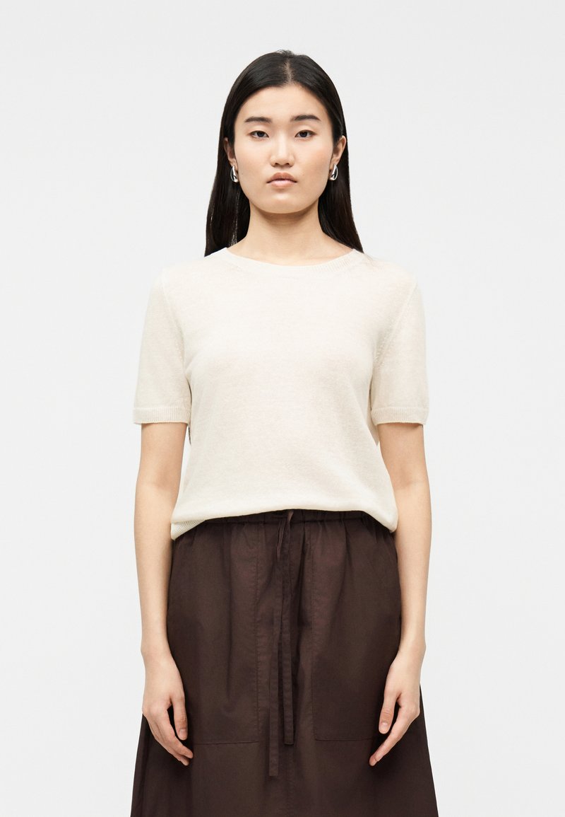 Femme aux longs cheveux noirs portant des boucles d'oreilles anneaux argentées, un pull crème à manches courtes, et une jupe taille haute marron foncé avec ceinture à cordon.