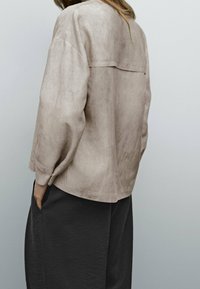 Chemise en daim beige à coupe décontractée, épaules tombantes et détail de yoke au dos, associée à un pantalon foncé, présentée sur un fond neutre.