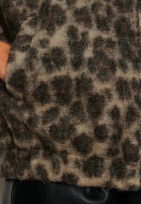 Tissu en polaire imprimé léopard beige et marron foncé, avec une surface douce et texturée. Présente un ourlet légèrement retroussé.