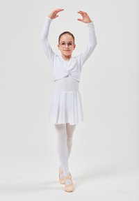Weißes Langarm-Tanzoutfit mit Knotendetail, gestuftem Rock, weißen Strumpfhosen und rosa Ballettschuhen. Das Model trägt eine Brille und posiert in einer Ballettpose.