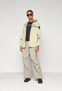 Šviesiai žalia striukė su pilkais akcentais, juodu logotipu, derinama su smėlio spalvos platėjančiomis kelnėmis. Po ja juodas marškinėlis, juodi batai ir akiniai nuo saulės.