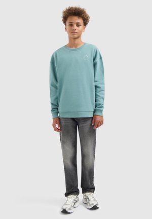 Teenager mit lockigem Haar, der einen türkisfarbenen Pullover, ausgewaschene schwarze Jeans und weiße Sneakers trägt, steht vor einem einfarbigen hellen Hintergrund.