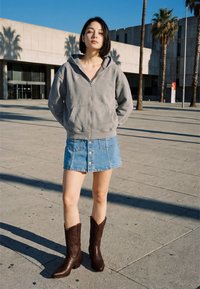 Fiatal nő áll kint egy szürke cipzáras kapucnis pulóverben, farmer gombos miniszoknyában és barna cowboy csizmában, mögötte pálmafák és egy épület látható.