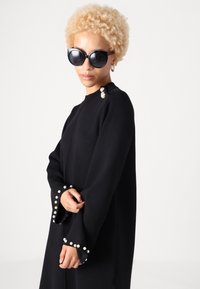 Čierne dlhé šaty s rukávmi, doplnené perlovými akcentmi na manžetách a pleciach, s okrúhlym výstrihom a voľným strihom. Hladká textúra látky.