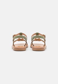 Des sandales beiges avec des sangles vertes, dotées d'une fermeture à boucle. Semelle plate avec des accents texturés et un design à dos ouvert. Style minimaliste.