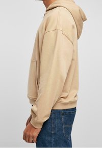 Felpa con cappuccio beige, con cordino, tasca a marsupio e polsini a costine. Indossata con jeans di denim blu scuro, che mostrano un fit rilassato.