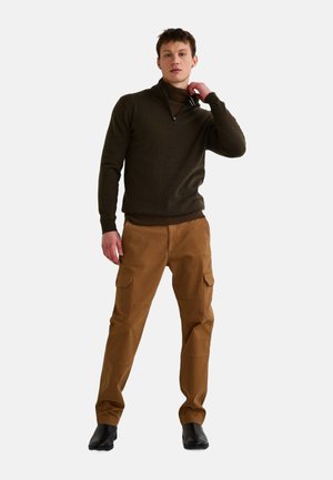 Pull zippé vert olive associé à un pantalon cargo beige avec de multiples poches et des chaussures noires. Textures lisses et design ajusté.
