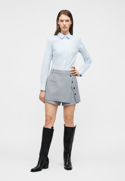 Γυναίκα που στέκεται φορώντας γαλάζιο πουκάμισο με κουμπιά, ασύμμετρο γκρι σορτς με κουμπιά και μαύρες μπότες μέχρι το γόνατο σε λευκό φόντο.