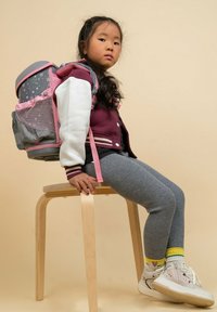 Rucksack mit pinken Akzenten und grauem Stoff mit Sternenmuster. Modell in einer burgunderfarbenen Jacke, grauen gerippten Leggings und weißen Turnschuhen. Sitzt auf einem Holzschemel.
