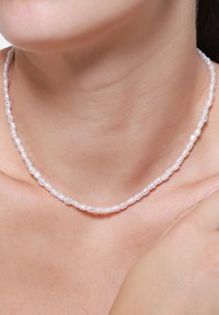 Biała perłowa naszyjnik wykonany z małych, owalnych koralików, gładka tekstura, równomiernie rozmieszczone, spoczywające na skórze z minimalnie widocznym detalem zapięcia.