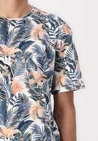 Tricou cu mânecă scurtă, având un model floral tropical multicolor cu accente albastre, piersici și verzi pe un fundal alb.
