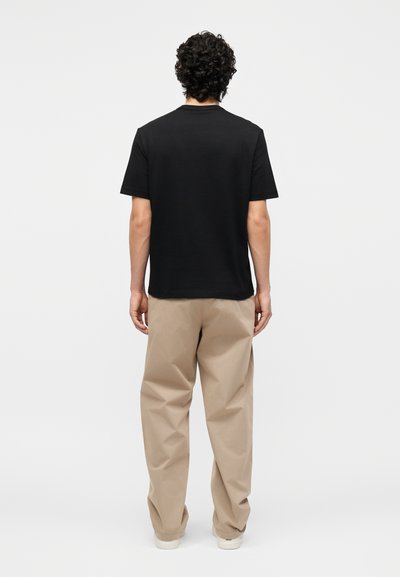 Άνδρας με σγουρά σκούρα μαλλιά στέκεται με την πλάτη, φορά ένα μονόχρωμο μαύρο μπλουζάκι και φαρδιά μπεζ παντελόνια σε λευκό φόντο.