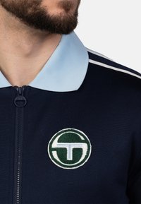 Marine blauwe zip-up jack met een lichtblauwe kraag en witte strepen. Bevat een ronde, groene en witte geborduurde logo op de linkerborst.